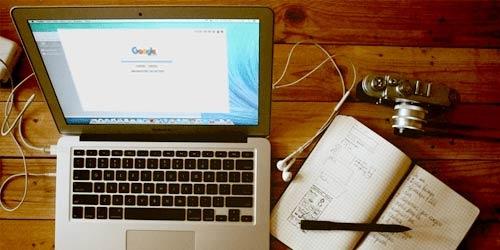 Das Bild zeigt einen mobilen Arbeitsplatz mit Notebook, Kamera und Notizbuch auf einem Holztisch. Auf dem aufgeklappten Notizbuch sieht man die Startseite von Google. Das Bild steht sinnbildlich dafür, dass deine Website mit einer sinnvollen Suchmaschinenoptimierung von überall her besser gefunden wird.